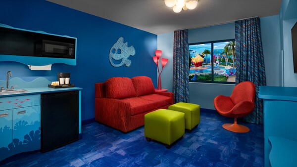 Interior de um quarto com piso laminado, uma cadeira estofada, 2 pufes, um sofá, uma TV de tela plana montada na parede, uma mesa lateral, uma cômoda, um micro-ondas, um frigobar, uma pia, cortinas e uma janela