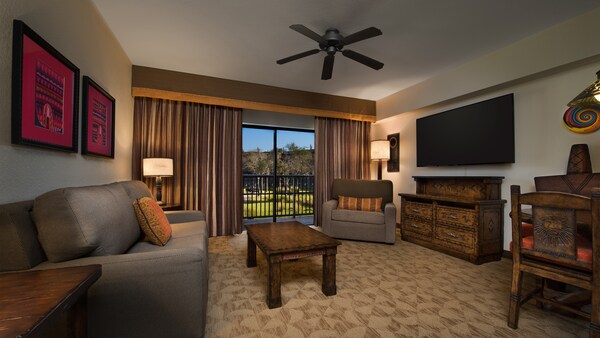 Una sala de estar decorada al estilo africano con una sofá, una mesa baja, cuadros, una silla, una cómoda un TV y un área de comidas