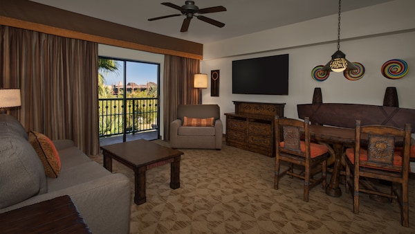 Una sala de estar decorada al estilo africano con una sofá, una mesa baja, una silla, lámparas, una cómoda, TV, una mesa de comedor y cuadros