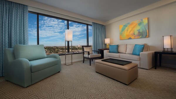 Un área de estar con un sofá, una otomana, un sofá de dos cuerpos, una silla, una lámpara, un cuadro y una ventana