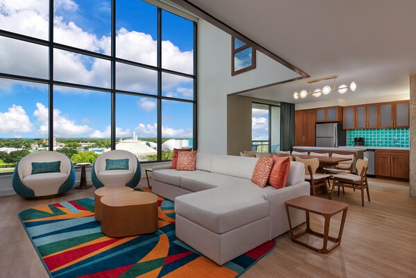Una sala de estar de una villa grande de lujo de 3 dormitorios con un estudio y vista al Parque Temático en Bay Lake Tower at Disney's Contemporary Resort, con un sofá, 2 sillas, una mesa de restaurante, una cocina y una ventana con vistas a Magic Kingdom Park