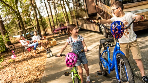 Dos niños caminando con sus bicicletas en la mano junto a una casa rodante estacionada mientras sus padres observan sentados en sillas plegables