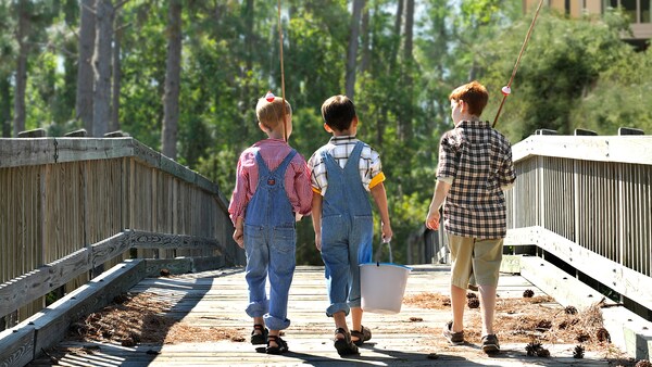 A trio of young male Guests clad in country wear cross a bridge while carrying fishing supplies