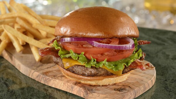 Una hamburguesa de carne de res con queso, tocineta, lechuga, rodajas de tomate, cebollas moradas y queso cheddar, cerca de una pila de papas fritas