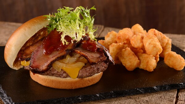 A hamburger topped with cheese, onions, a meat patty, sliced brisket and lettuce next to a pile of tater tots