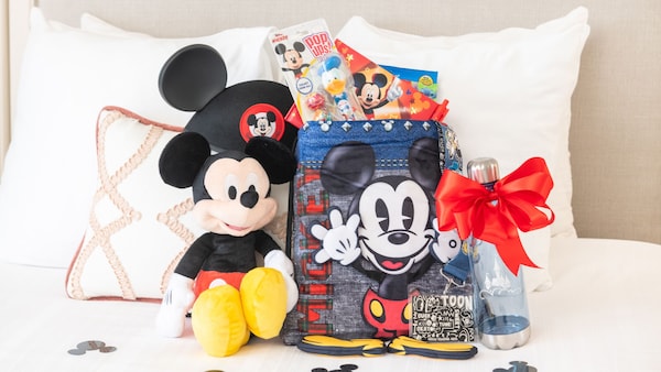 A Mickey Mouse plush, a water bottle, a Mickey Mouse ear hat and other Mickey Mouse merchandise displayed on a bed against pillows	