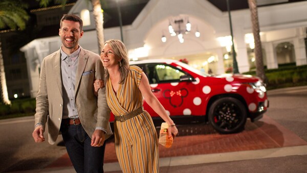 A man and woman dressed in semi formal attire, walking past a Minnie Van SUV at the entrance to a Disney Resort hotel