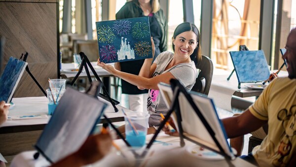 A woman holds up her painting of Cinderella Castle surrounded by fireworks during a painting class 
