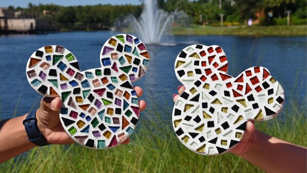 Deux personnes tiennent deux oeuvres d’art en mosaïque qui représentent les têtes de Mickey et de Mini devant un lac avec une fontaine