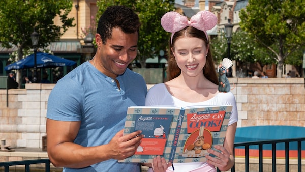 A couple at Epcot posing with an augmented reality overlay of a cookbook and Remy from Ratatouille