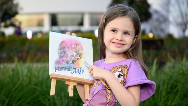 Una niña al aire libre sosteniendo un pequeño caballete con una pintura con el texto 'Festival of the Arts'
