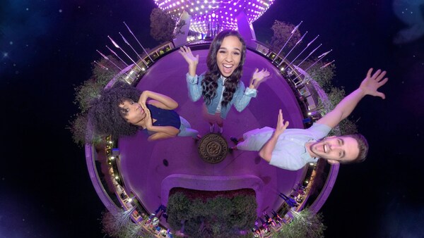 Tres Visitantes posan frente a una representación esférica estilizada de EPCOT por la noche y las palabras 'Epcot International Festival of the Arts'