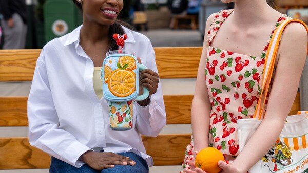 Two Guests displaying fruit themed apparel, including a dress, travel cup and tote bag, while sitting on a bench