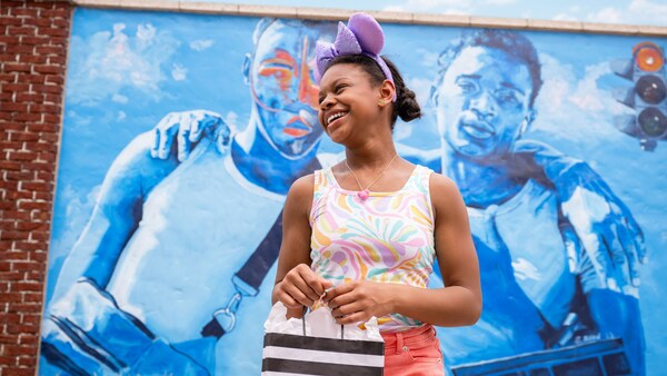 A young woman in a Minnie Ear Headband holds a gift bag near a mural of 2 young men, arm in arm