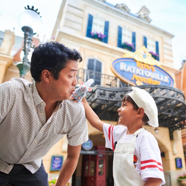 A young Guest holding a Remy plushie up to his father’s shoulder in front of Remy’s Ratatouille Adventure at Epcot