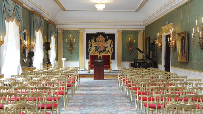 Un majestuoso salón de recepciones con sillas, un piano y un podio con un letrero que dice “Hillsborough Castle”