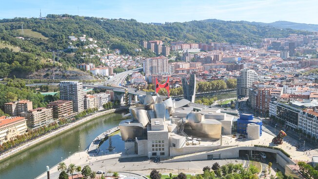 El paisaje urbano de Bilbao revela montañas verdes, el museo curvilíneo de Guggenheim, un río y una serie de edificios de varios pisos