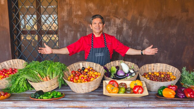 A man smiles while his hands extend over baskets filled with fresh vegetables