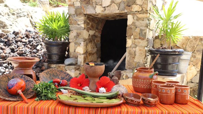 A table with herbs and Mexican pottery next to the entrance of a small stone hut