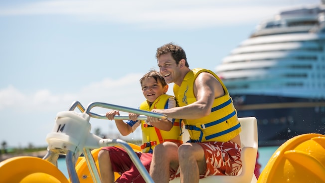 A father and son steer an Aqua Cycle Water Trike with a Disney Cruise Line ship behind them A father and son steer an Aqua Cycle Water Trike with a Disney Cruise Line ship behind them
