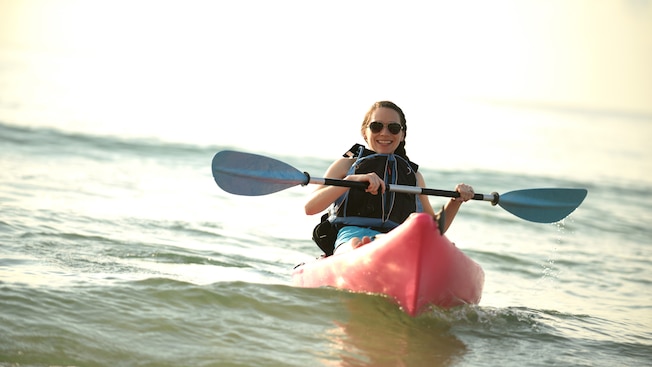 Una mujer con lentes de sol y un chaleco salvavidas sonríe mientras rema sobre un kayak en el mar Una mujer con lentes de sol y un chaleco salvavidas sonríe mientras rema sobre un kayak en el mar