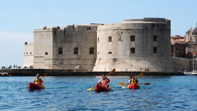 3 praticantes de caiaque remando pela água em frente à fortificação de pedra de Dubrovnik 3 praticantes de caiaque remando pela água em frente à fortificação de pedra de Dubrovnik