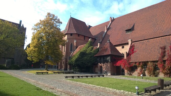 The courtyard of a large brick castle reveals trees, lawn and benches