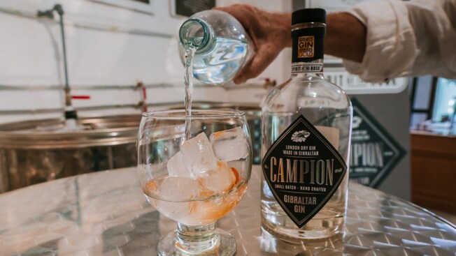 A bottle of gin next to a hand pouring a liquid into an oversized snifter glass containing ice cubes