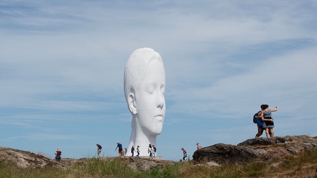 Visitantes caminhando e tirando selfies em um ambiente natural acidentado com uma grande escultura da cabeça de uma mulher