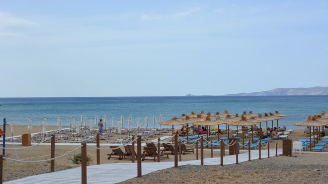 A public beach facing the Aegean Sea, featuring a wooden walkway leading to an area with a large number of sunbeds and umbrellas