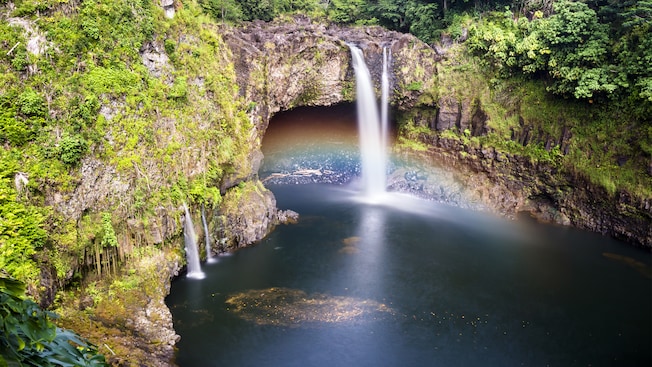 A waterfall flows into a lagoon from a source above the entrance to a cave carved into a rock wall overgrown with plants