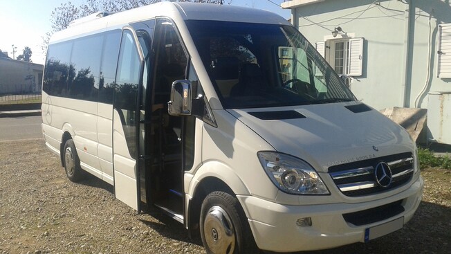 A minibus with its door open and a Mercedes Benz logo on the front is parked in front of a building