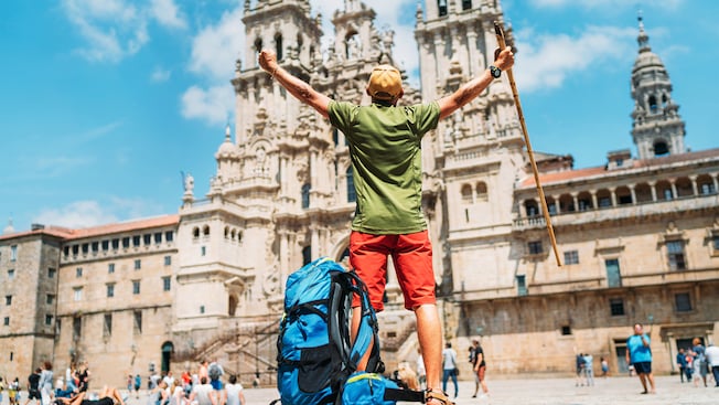 A man with a walking stick and backpack raising his hands in triumph in front of the Gothic cathedral of Santiago de Compostela A man with a walking stick and backpack raising his hands in triumph in front of the Gothic cathedral of Santiago de Compostela