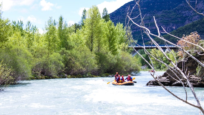 A group of 5 outfitted in helmets and lifejackets rafting on a river lined with trees A group of 5 outfitted in helmets and lifejackets rafting on a river lined with trees