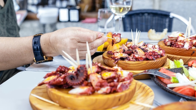 A hand reaching for a piece of grilled calamari on a table with a glass of wine and several plates of appetizers A hand reaching for a piece of grilled calamari on a table with a glass of wine and several plates of appetizers