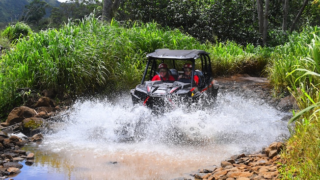 2 people ride a UTV through a puddle near a forest