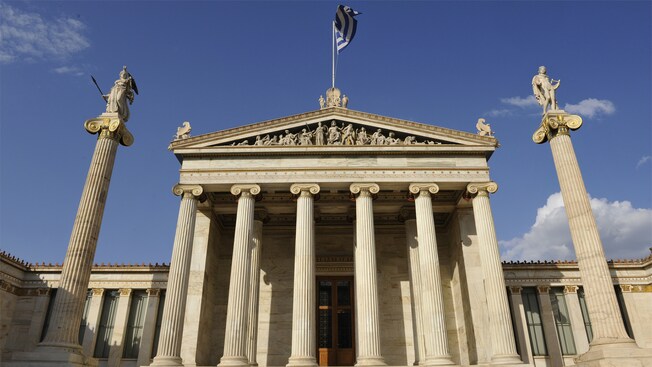 The Academy of Athens features a Greek flag flying over a monumental building with columns, a cornice and statues