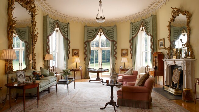The interior of a room containing a fireplace, large mirrors, antiques, upholstered chairs, a couch, tables and books