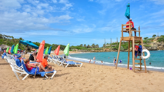 People lounging shoreside on a sandy beach while a lifeguard watches families playing in the water People lounging shoreside on a sandy beach while a lifeguard watches families playing in the water