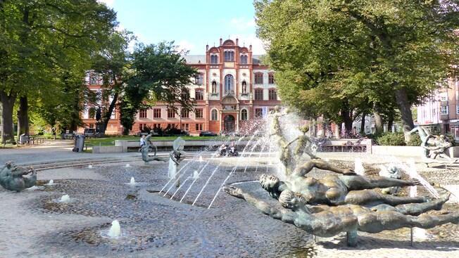 Árvores maduras e uma grande fonte com estátuas de bronze em frente a um edifício monumental