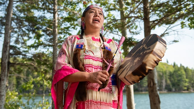 A woman in the traditional attire of an indigenous community, singing and playing a drum by a lake A woman in the traditional attire of an indigenous community, singing and playing a drum by a lake