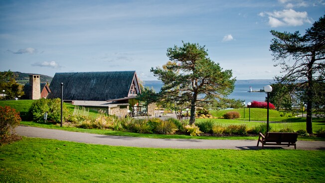 A modern A-frame building on verdant grounds overlooking a body of water and an outcropping with a lighthouse