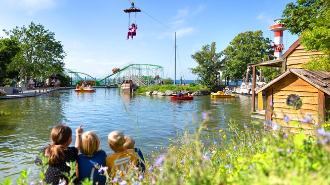 A busy amusement park with a roller coaster, a lake with a boating attraction and a person gliding overhead in a ski lift