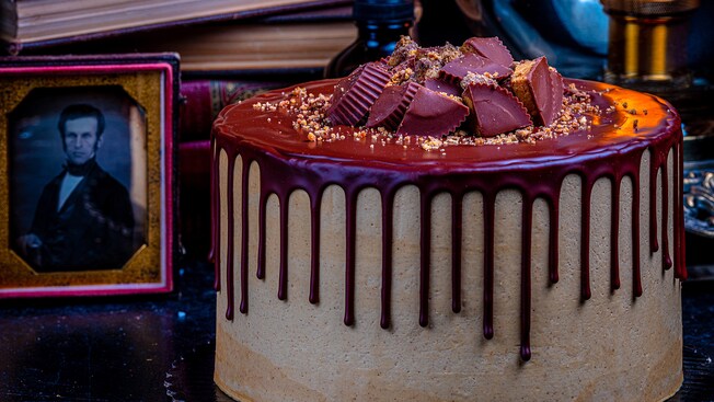 A Peanut Butter Cake topped with chunks of Peanut Butter Cups with chocolate ganache pouring over the sides