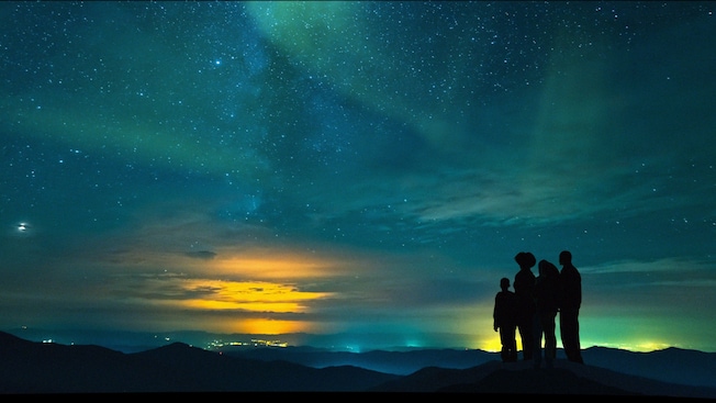 Family stands on a mountain looking at a beautiful blue and yellow night sky