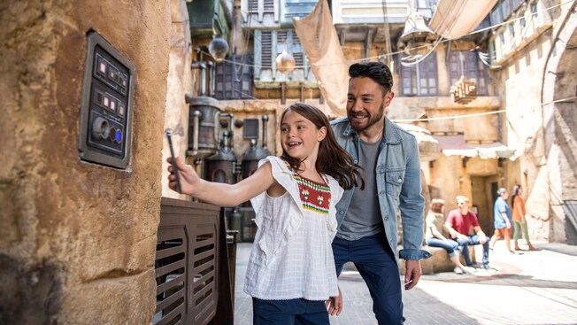 A dad watches his daughter hold a cell phone up to a computer display imbedded in a stone wall