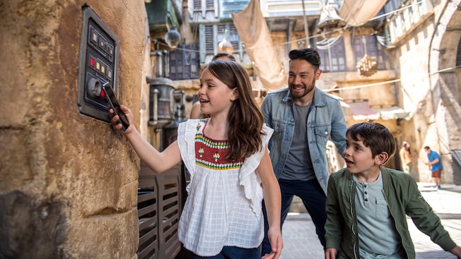 A girl smiles and holds a cell phone to a computer display while her parents and brother watch