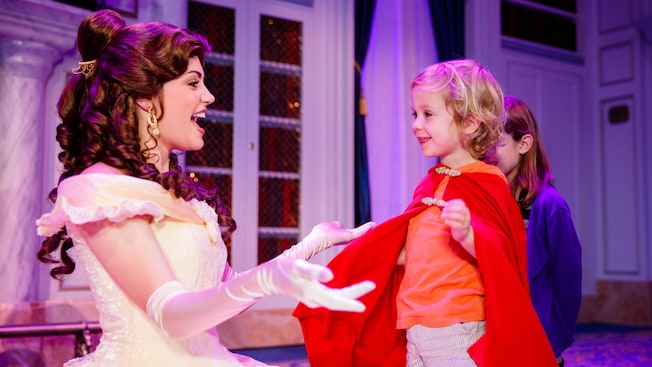 Belle kneels down to a hug a child wearing a cape
