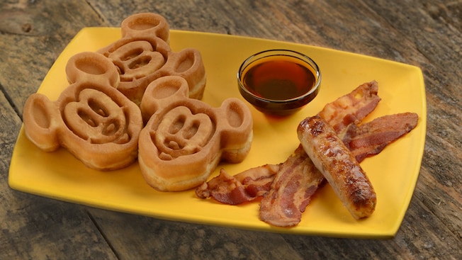 Three waffles in the shape of Mickey Mouse, a bowl of syrup, bacon and sausage on a plate