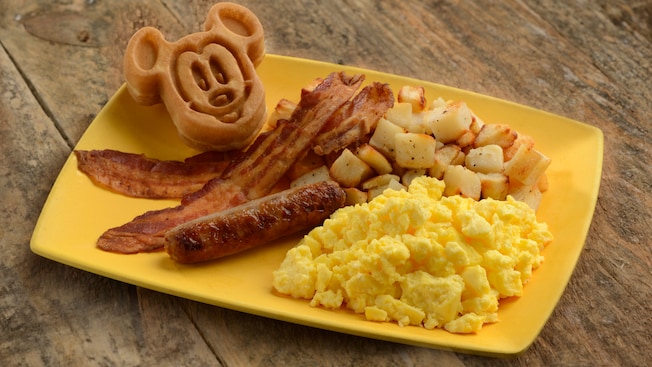 A waffle in the shape of Mickey Mouse, bacon, sausage, scrambled eggs and potatoes on a plate